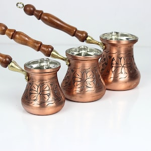 May include: Three copper coffee pots with wooden handles and floral etched designs. The pots have a shiny copper finish with silver interiors. The handles are a dark brown color. The pots are arranged in a row, with the smallest pot in the front.