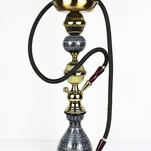 Puede incluir: Una cachimba decorativa con un cuenco de terracota, detalles en latón y una manguera negra. La base es negra con un patrón blanco. La cachimba mide aproximadamente 61 cm de altura.
