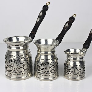 Peut inclure: Un ensemble de trois cafetières en métal argenté avec des manches en bois foncé. Chaque pot présente un bord large et évasé, un col étroit et un corps cylindrique orné de motifs floraux gravés complexes et d'une petite boucle de suspension.