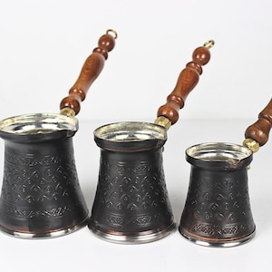 May include: Three dark copper coffee pots of varying sizes, each featuring a tapered body with an embossed geometric pattern, a silver-toned interior, and a long, turned wooden handle with a small metal loop at the end for hanging storage.