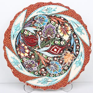 May include: A decorative ceramic plate with a scalloped edge. The plate features a detailed floral design in red, blue, yellow, and purple against a black background. The rim is a reddish-brown color with small circular patterns.