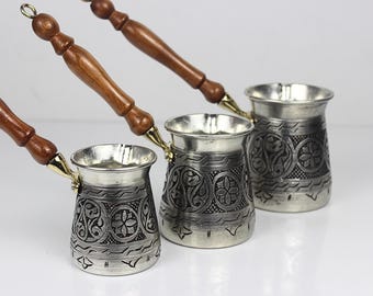May include: Three silver-colored Turkish coffee pots with ornate floral and geometric patterns. Each pot has a wooden handle and a gold-colored accent where the handle connects to the pot. The pots are displayed on a white surface.