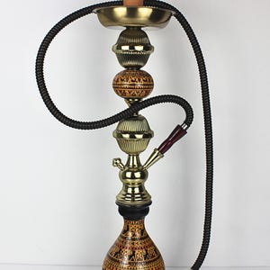 May include: An ornate hookah with a terracotta bowl, brass accents, and a patterned glass base. The hookah has a black hose and a wooden handle. The base features a detailed, hand-painted design in red, black, and gold.