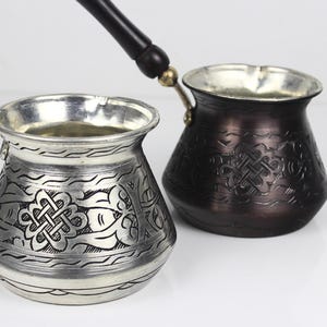 Peut inclure: Deux cafetières turques décoratives. L'une est de couleur argentée avec des motifs floraux gravés et une poignée en bois noir. L'autre est marron foncé avec des motifs gravés similaires et une poignée noire.