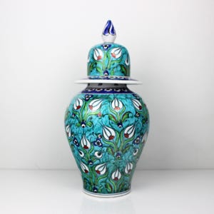 May include: A ceramic jar with a lid, featuring a turquoise background and a floral pattern in white, red, and green. The jar has a bulbous shape and a matching lid with a decorative finial. The design is reminiscent of traditional Turkish pottery.