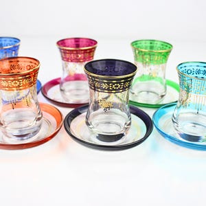 Op de afbeelding: Set van zes kleurrijke glazen theekopjes met bijpassende schotels. Elke kop heeft een andere gekleurde rand en schotel, waaronder blauw, rood, groen, paars, oranje en turquoise. De randen zijn versierd met gouden bloempatronen.