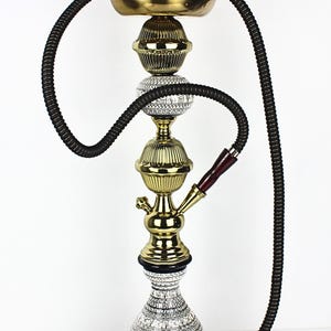 Puede incluir: Una cachimba ornamentada con un diseño decorativo. La cachimba presenta un cuerpo de metal dorado, una manguera negra y un cuenco de cerámica. La base es blanca con un patrón negro. La cachimba está diseñada para fumar.