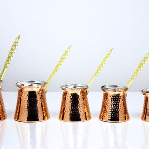 May include: A set of five graduated copper coffee pots with a hammered exterior finish and a metallic silver-toned interior lining. Each pot features a long, slender, textured gold-colored metal handle attached to the side of the vessel.
