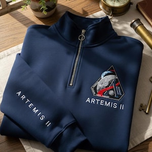 May include: A navy blue Artemis II quarter-zip sweatshirt. The sweatshirt has a silver zipper, a triangular patch with a space-themed design, and the words "ARTEMIS II" printed on the sleeve and below the patch. The sweatshirt is made of a soft material.