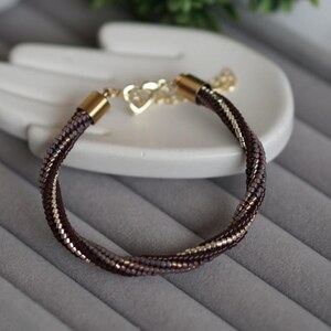 Puede incluir: Una pulsera de cuentas trenzadas con cuentas de semillas de color morado oscuro y oro metálico. La pulsera tiene terminales de metal dorado y un dije en forma de corazón dorado unido a una cadena extensora con un cierre de mosquetón.