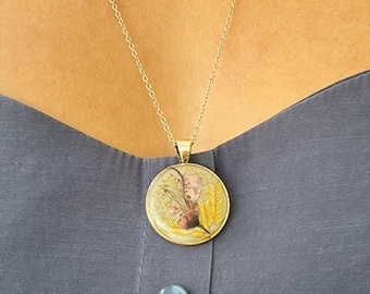 Puede incluir: Un collar con colgante dorado con un colgante redondo y detallado. El colgante presenta una ilustración colorida con tonos amarillos, rosas y marrones. El collar se lleva sobre una parte superior azul grisáceo.