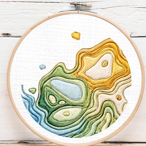 May include: Embroidered landscape art in a wooden hoop. The design features a topographic map with shades of blue, green, and yellow. The text "Digital Download (PDF)" is in a green circle in the upper left corner.