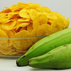 Può includere: Una ciotola di vetro piena di patatine di platano giallo dorato è accanto a due platani verdi. Le patatine sono sottili e croccanti, e i platani sono lunghi e curvi. Il cibo è su una superficie bianca.
