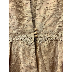 May include: Close-up of a beige lace jacket with a floral pattern. The jacket features three pearl-like buttons and decorative floral trim. The fabric has a delicate, textured appearance, suitable for formal occasions.