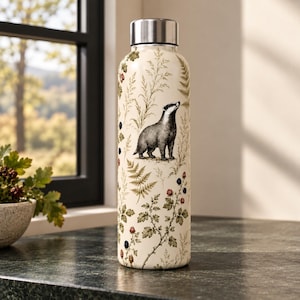 May include: A cylindrical water bottle with a cream-coloured background featuring a detailed illustration of a badger standing among green ferns, tall grasses, and small clusters of red and black berries, topped with a metallic silver screw-top lid.
