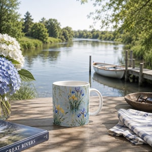 Può includere: Una tazza in ceramica bianca con una stampa dettagliata in stile acquerello di iris blu e gialli che crescono nell'acqua. La tazza ha un corpo cilindrico liscio e un manico curvo, con un motivo botanico su uno sfondo azzurro.
