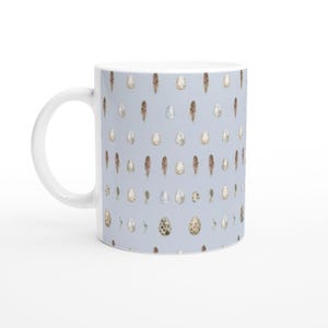 Puede incluir: Taza de cerámica blanca con un patrón repetido de plumas y huevos moteados en acuarela sobre un fondo azul claro. La taza tiene un asa y un interior blancos. El diseño tiene un tema de la naturaleza.