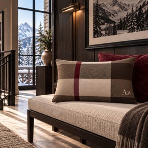May include: A rectangular throw pillow featuring a brown, cream, and dark red plaid pattern. The pillow has the letters A&W embroidered in white on the bottom right corner. It is positioned on a light-colored bench with a dark brown wooden frame.