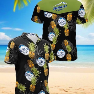 Puede incluir: Una camisa de manga corta con botones, fondo negro y un patrón repetitivo de piñas con gafas de sol y el logotipo de Busch Light. La parte posterior tiene un canesú de color verde oliva sólido con el logotipo de Busch Light impreso.