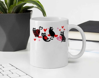 May include: White ceramic mug featuring a repeating pattern of black cat illustrations with red hearts and pink stars. The cats are holding hearts or yarn. The mug has a large handle and is set against a neutral background.