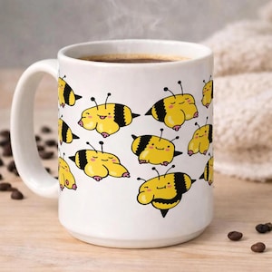 Puede incluir: Taza de café de cerámica blanca con un patrón repetido de abejas de dibujos animados con cuerpos amarillos, rayas negras y caras sonrientes. La taza está llena de café oscuro y tiene un asa blanca. Granos de café esparcidos.