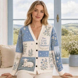 Puede incluir: Un cárdigan de manga larga con botones y diseño de patchwork en tonos azul y crema. El tejido de punto muestra patrones florales, motivos de estrellas y el gráfico de un cordero blanco. El texto The Lord Is My Shepherd está impreso al frente.