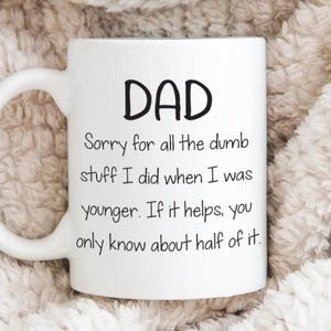 May include: White ceramic mug with the word "DAD" in bold black letters. Below, the mug has a humorous message: "Sorry for all the dumb stuff I did when I was younger. If it helps, you only know about half of it."