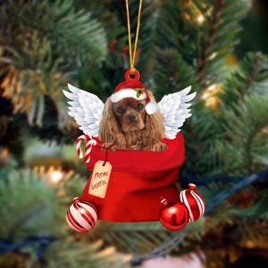 May include: A Christmas ornament featuring a brown Cavalier King Charles Spaniel wearing a Santa hat with white angel wings, peeking out of a red gift sack. The sack has a tag labeled From Santa and is decorated with red and white striped ornaments.
