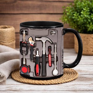 Puede incluir: Taza de café negra con un diseño de caja de herramientas gris que muestra varias herramientas, como alicates, una llave inglesa, un martillo y un destornillador. La taza tiene un asa y un borde negros. Las herramientas son rojas, negras y plateadas.