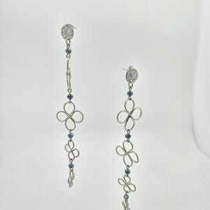 May include: A pair of silver-toned dangle earrings. Each earring features a small, textured disc stud, connected to a chain with blue beads and openwork floral designs. The earrings have a delicate, feminine aesthetic.