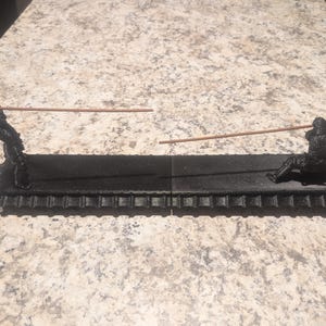 Puede incluir: Porta incienso negro con dos figuras, una de pie y otra reclinada, sosteniendo varillas de incienso. El soporte tiene un borde decorativo festoneado. Las varillas de incienso son de color marrón claro.