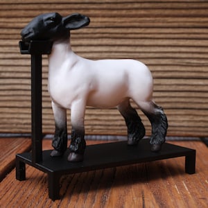 May include: A white and black sheep figurine stands on a black metal display stand. The sheep has a black head and legs, with a white body. The stand is rectangular with four legs and a vertical support.