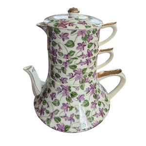 May include: A stacked teapot set with a floral violet pattern. The set includes a teapot, two cups, and a lid. The handles and accents are gold-colored. The background is white.