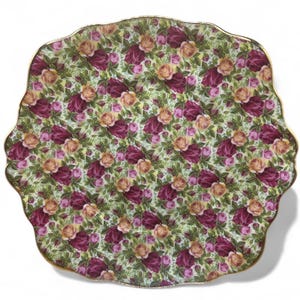 May include: A decorative, scalloped-edge plate with a floral pattern. The plate features a repeating design of burgundy and peach roses with green leaves on a light green background. The rim is trimmed with a gold border. The plate is likely made of ceramic or porcelain.