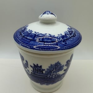 May include: A white ceramic sugar bowl with a lid, featuring a blue transferware pattern depicting a landscape with pagodas, trees, and bridges. The lid has a small knob handle, and the body of the bowl has a tapered shape with a smooth finish.