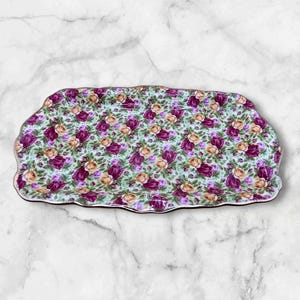 May include: Rectangular ceramic serving tray with a floral pattern. The tray features a light green background with a repeating pattern of purple and peach-colored roses. The edges of the tray are scalloped and trimmed with a gold border.