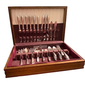 May include: A wooden storage chest with a hinged lid containing a set of silver-toned flatware. The interior features a cream-colored pleated fabric lining and a dark maroon velvet base with individual slots for knives, forks, and spoons.