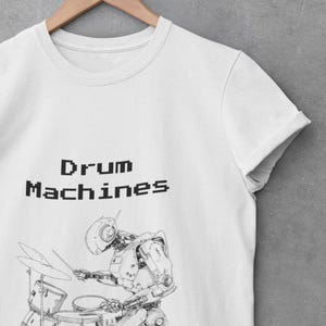 Puede incluir: Camiseta blanca con una ilustración en blanco y negro de un robot tocando la batería. El texto "Drum Machines Have No Soul" está impreso encima y debajo del gráfico del robot. La camiseta está colgada en una percha de madera.