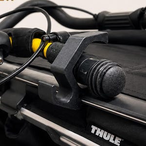 Op de afbeelding: Close-up van een rode en zwarte fietsaanhangerbevestiging. De bevestiging heeft de tekst "TRANSPORTHAKEN FÜR THULE ANHÄNGER" en "SICHER. STABIL. DURCHDACHT." De bevestiging is ontworpen voor een Thule-aanhanger.
