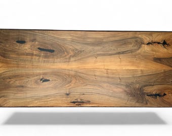 May include: A rectangular wooden table top with a natural wood grain pattern. The wood has a mix of light and dark brown tones, with dark knots and streaks. The edges are dark brown. The table top is likely for a dining or coffee table.