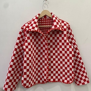 May include: A red and white checkered jacket hangs on a wooden hanger. The jacket has a collared neckline and a button-up front. The pattern is a classic checkerboard design, with red and white squares. The jacket is made of a quilted material.