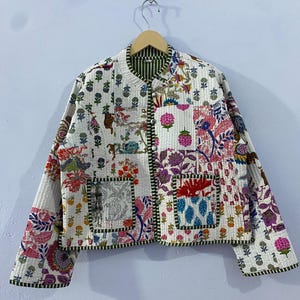May include: A quilted jacket with a patchwork design featuring floral and animal motifs in shades of pink, purple, and green on a white background. The jacket has a round collar and a button-up front.