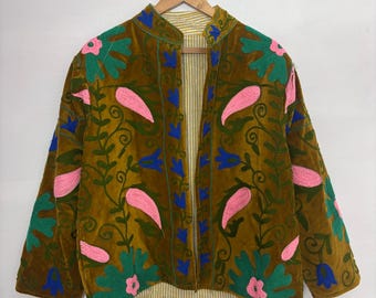Könnte beinhalten: Braune Samtjacke mit floralen Stickereien in Rosa, Grün und Blau. Die Jacke hat einen Stehkragen und ist vorne offen, wodurch ein gestreiftes Futter sichtbar wird. Sie hängt an einem Holzkleiderbügel.