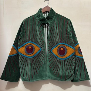 Op de afbeelding: Groene fluwelen jas met geborduurde oogontwerpen in goud, blauwgroen en bordeaux. De jas heeft een hoge kraag en een ritssluiting. De mouwen zijn wijd en de jas hangt aan een houten hanger.