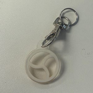 May include: A clear, circular keychain charm with a three-part design, attached to a silver-coloured clasp and keyring. The charm is made of a translucent material, and the metal hardware is shiny.
