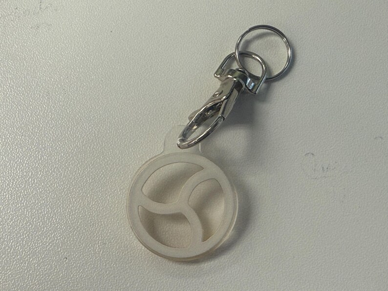 May include: A clear, circular keychain charm with a three-part design, attached to a silver-coloured clasp and keyring. The charm is made of a translucent material, and the metal hardware is shiny.