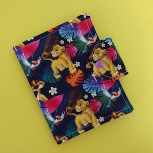 May include: A rectangular fabric pouch and a smaller matching coin purse featuring a dark blue background with a colorful jungle print. The pattern includes illustrations of lion cubs, a meerkat, and a bird surrounded by vibrant tropical leaves.