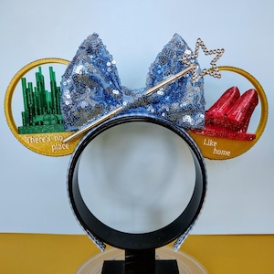 May include: A themed headband featuring a blue sequined bow, a gold wand with a star, and two circular designs. One design shows a green city, the other shows red shoes. Text on the headband reads "There's no place" and "like home".
