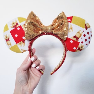May include: A pair of Minnie Mouse ears with a gold sequined bow and a red and gold headband. The ears feature a patchwork design with Winnie the Pooh illustrations and red polka dots.