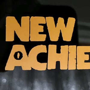 Könnte beinhalten: Nahaufnahme einer dunklen Oberfläche mit den Worten "NEW ACHIEVEMENT!" in einer kräftigen, orangefarbenen Schrift. Der Text ist zentriert und nimmt den größten Teil des Bildes ein. Das Ausrufezeichen ist deutlich sichtbar. Der Hintergrund ist unscharf.
